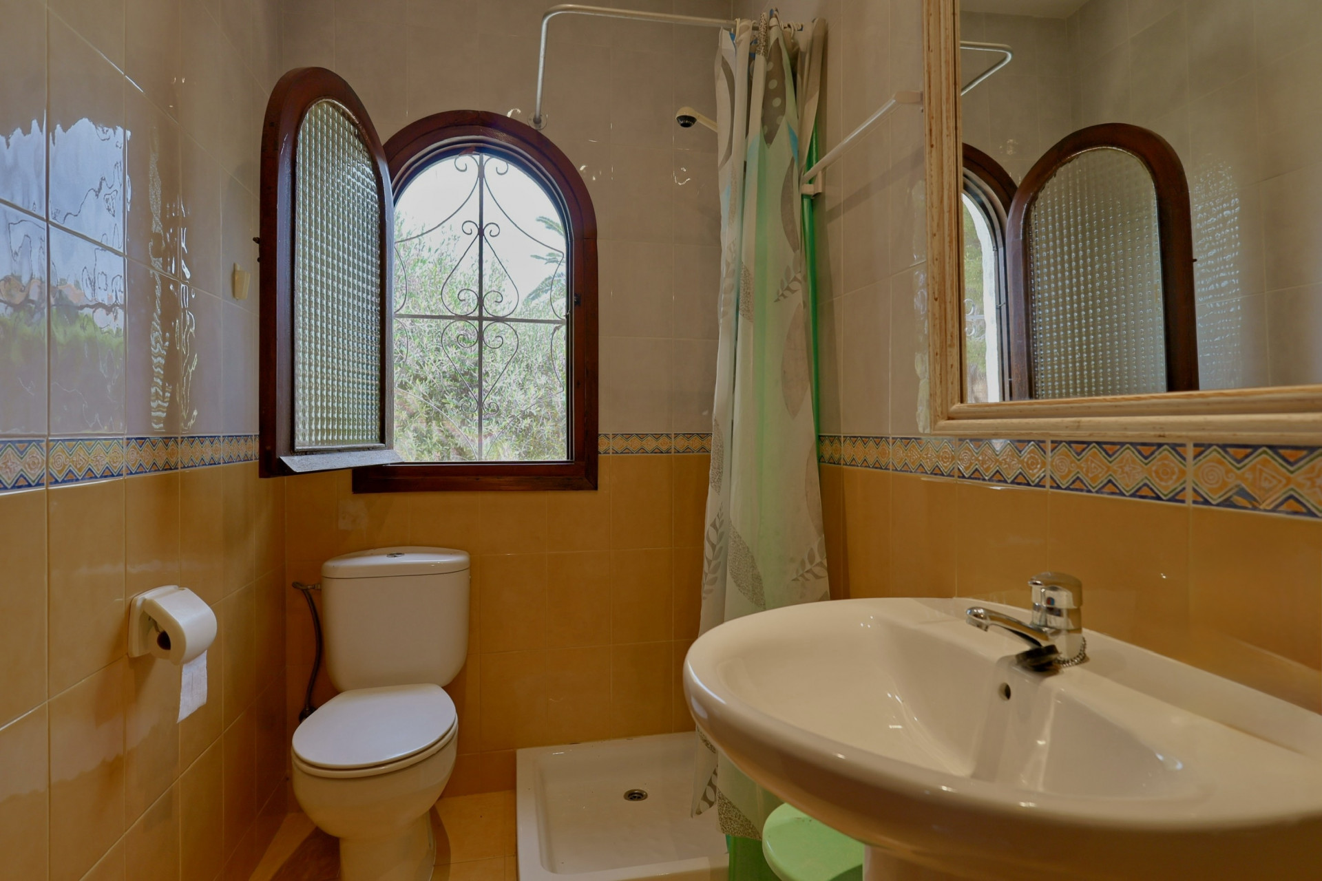 Badkamer met boogvormig houten raam, gestructureerd glas en smeedijzeren traliewerk. Tweekleurige tegelwanden met decoratieve rand, wit keramisch toilet en wastafel.