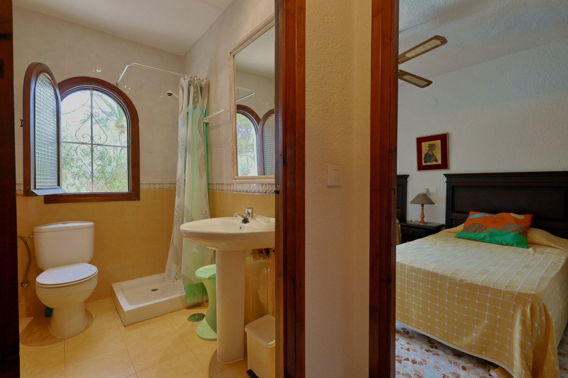 Badkamer met douche, toilet en wastafel op zuil, voorzien van een boogvormig houten raam met uitzicht op groen. Een glimp van een aangrenzende slaapkamer is zichtbaar.