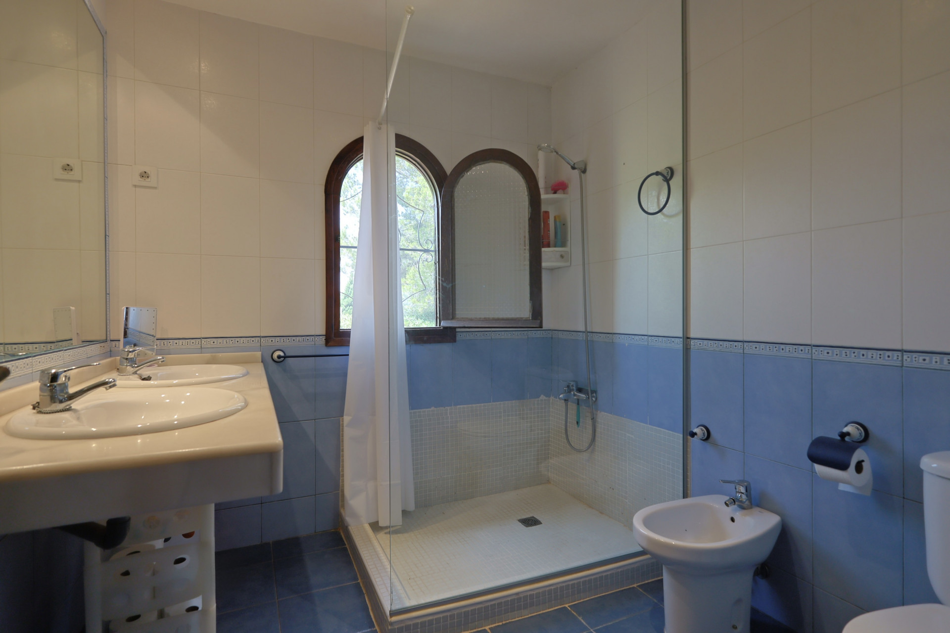 Badkamer met dubbele wastafel en licht aanrechtblad, wit-blauwe tegelwanden en inloopdouche met glazen scherm. Twee boogvormige houten ramen zorgen voor natuurlijk licht.