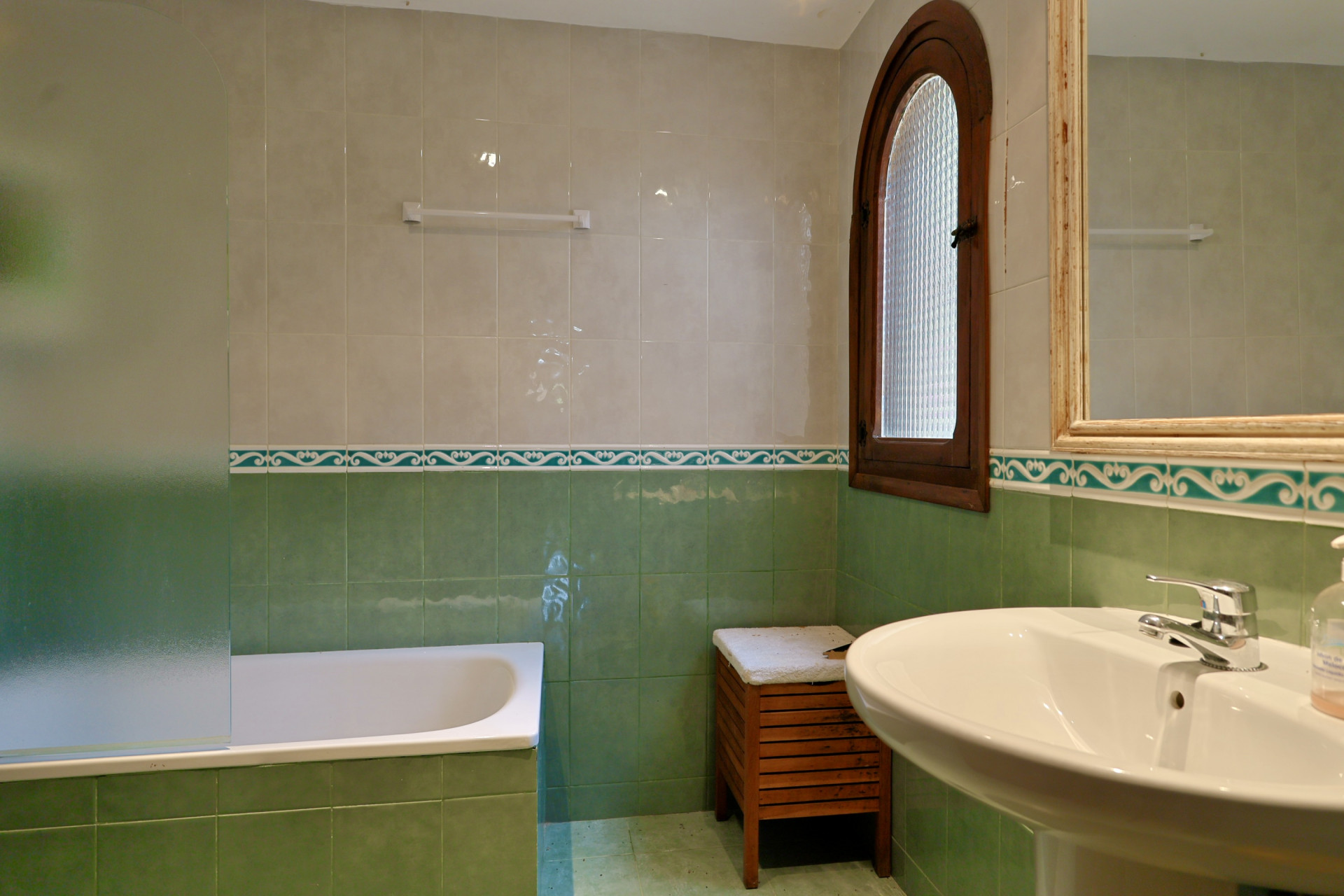 Badkamer met groen-beige tegels, ligbad met matglazen scherm en voetstukwastafel. Boograam van hout met gestructureerd glas biedt natuurlijk licht.