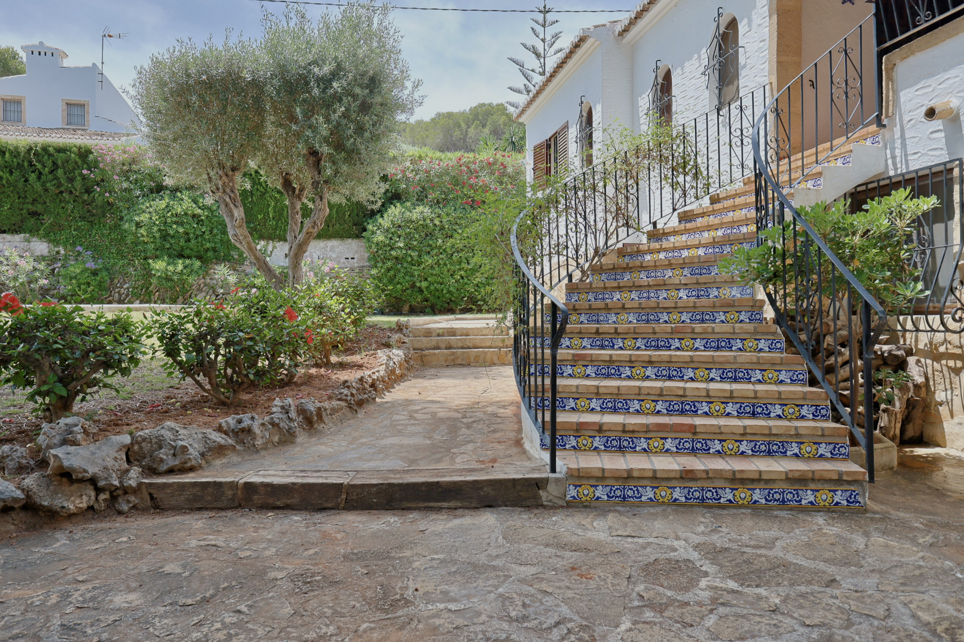 Buitentrap van baksteen met decoratieve blauw-gele tegels en smeedijzeren balustrade, geflankeerd door een mediterrane tuin met olijfboom.