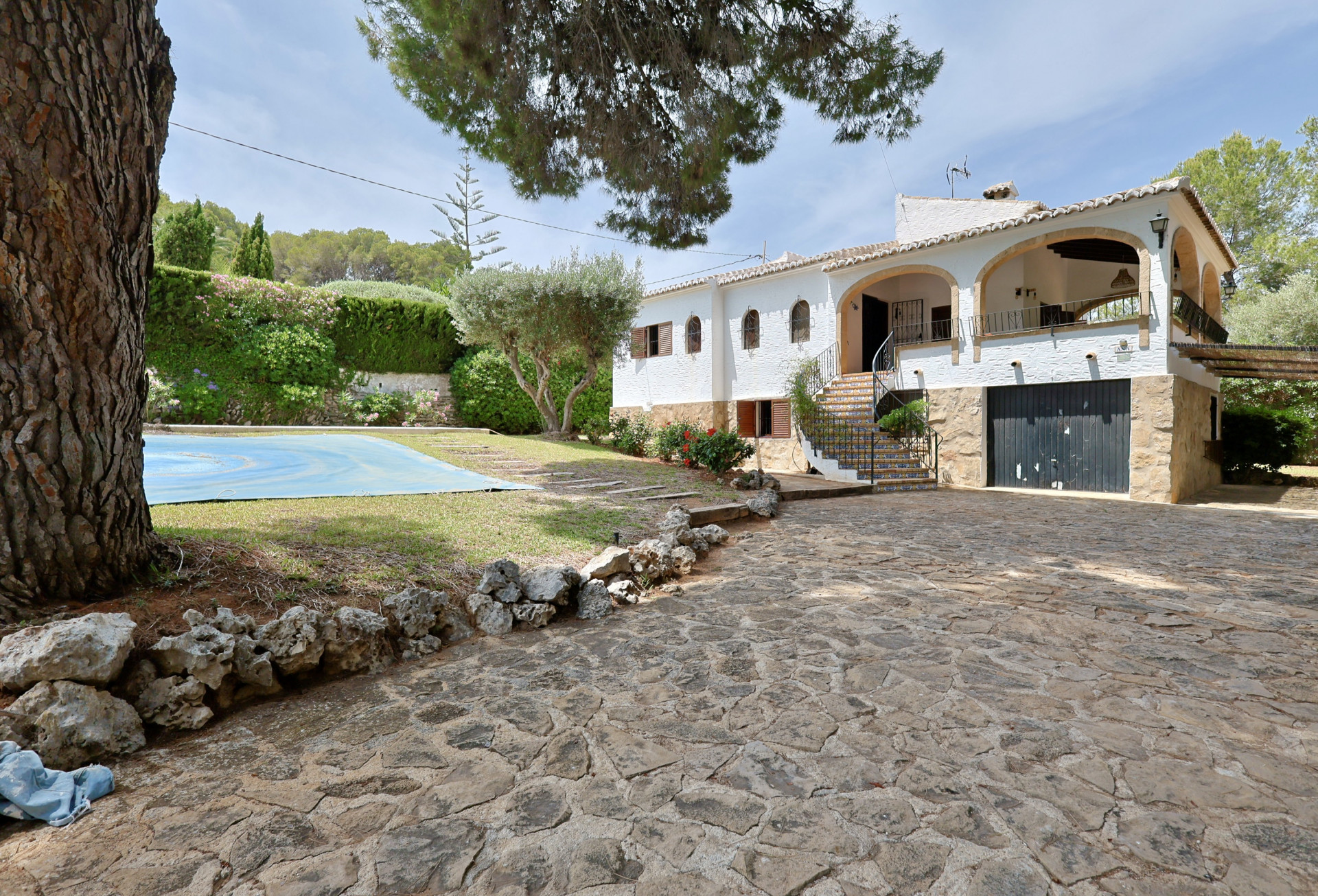 Buitenzijde van de villa met ruime geplaveide oprit, witte gevel met bogen en de hoofdtrap. Het afgedekte zwembad en mediterrane beplanting zijn zichtbaar.