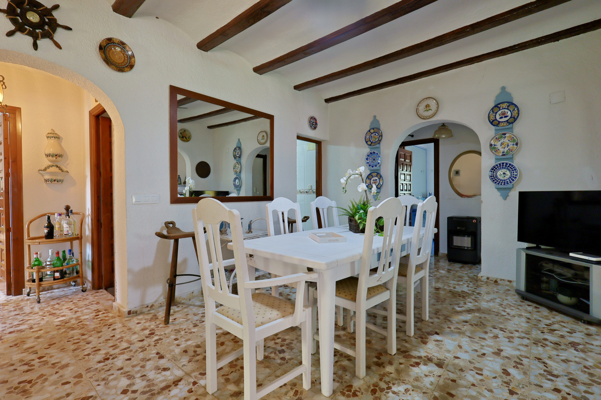 Eetkamer met witte houten tafel en stoelen, een terrazzo vloer en donkere plafondbalken. Decoratieve bogen en wandborden sieren de ruimte.