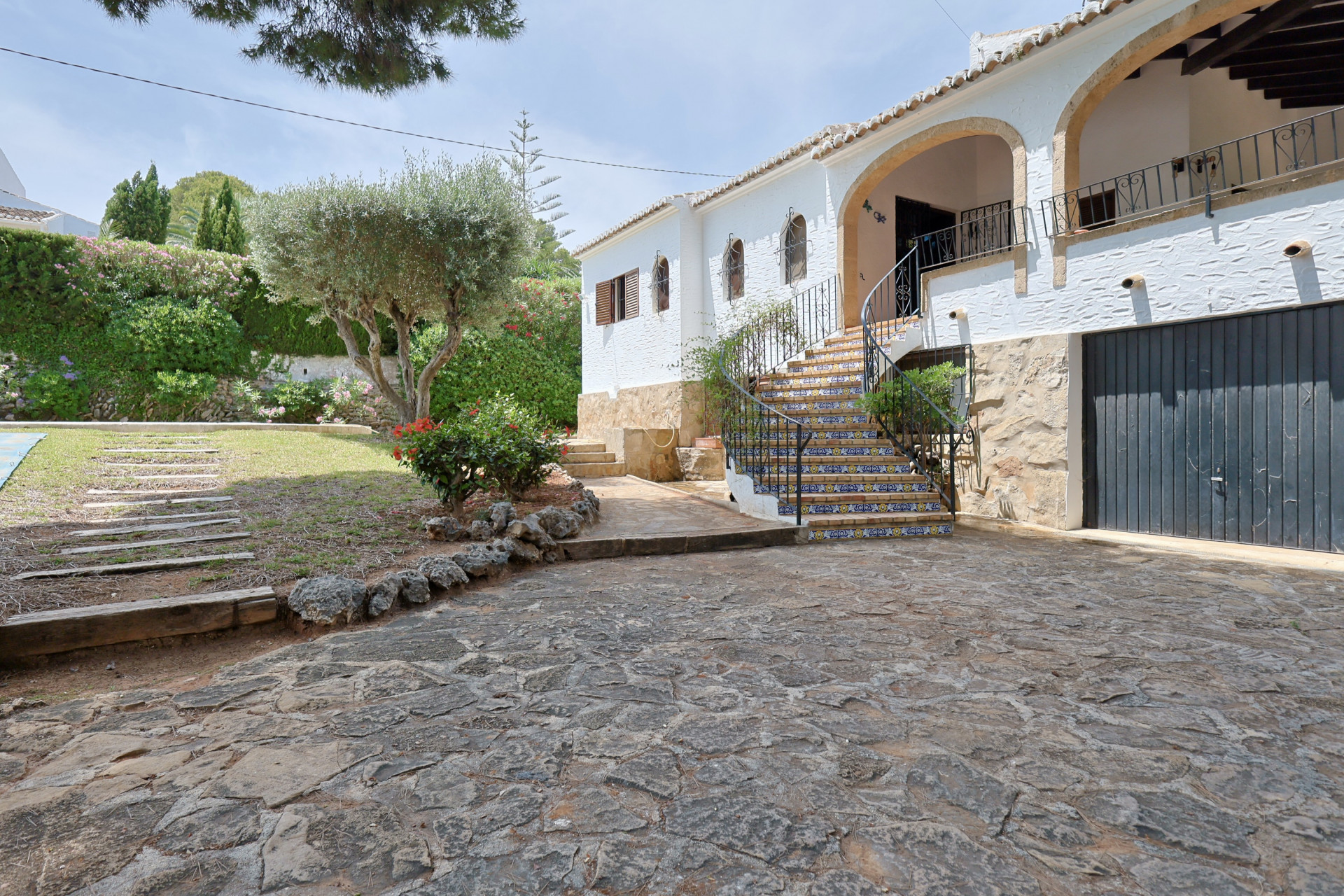 Exterieur van de villa met stenen oprit, een gebogen mozaïektrap en smeedijzeren balustrade. De mediterrane tuin omvat een olijfboom en een gazon naast de witte gevel.