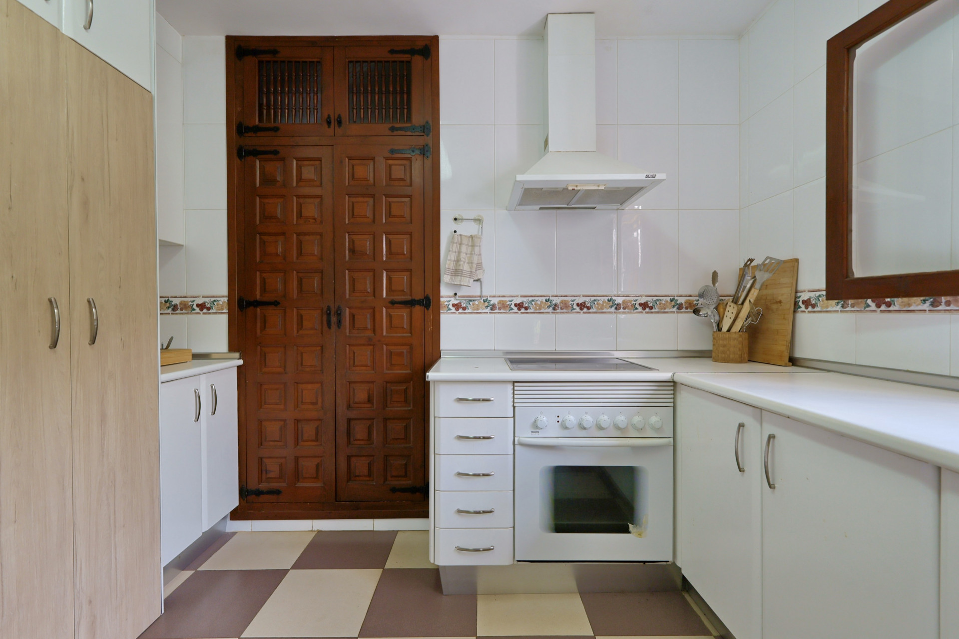 Functionele keuken met ingebouwde oven, afzuigkap en witte tegelwanden. Een opvallende grote houten kast met paneeldeuren en traditioneel beslag staat op een geblokte vloer.