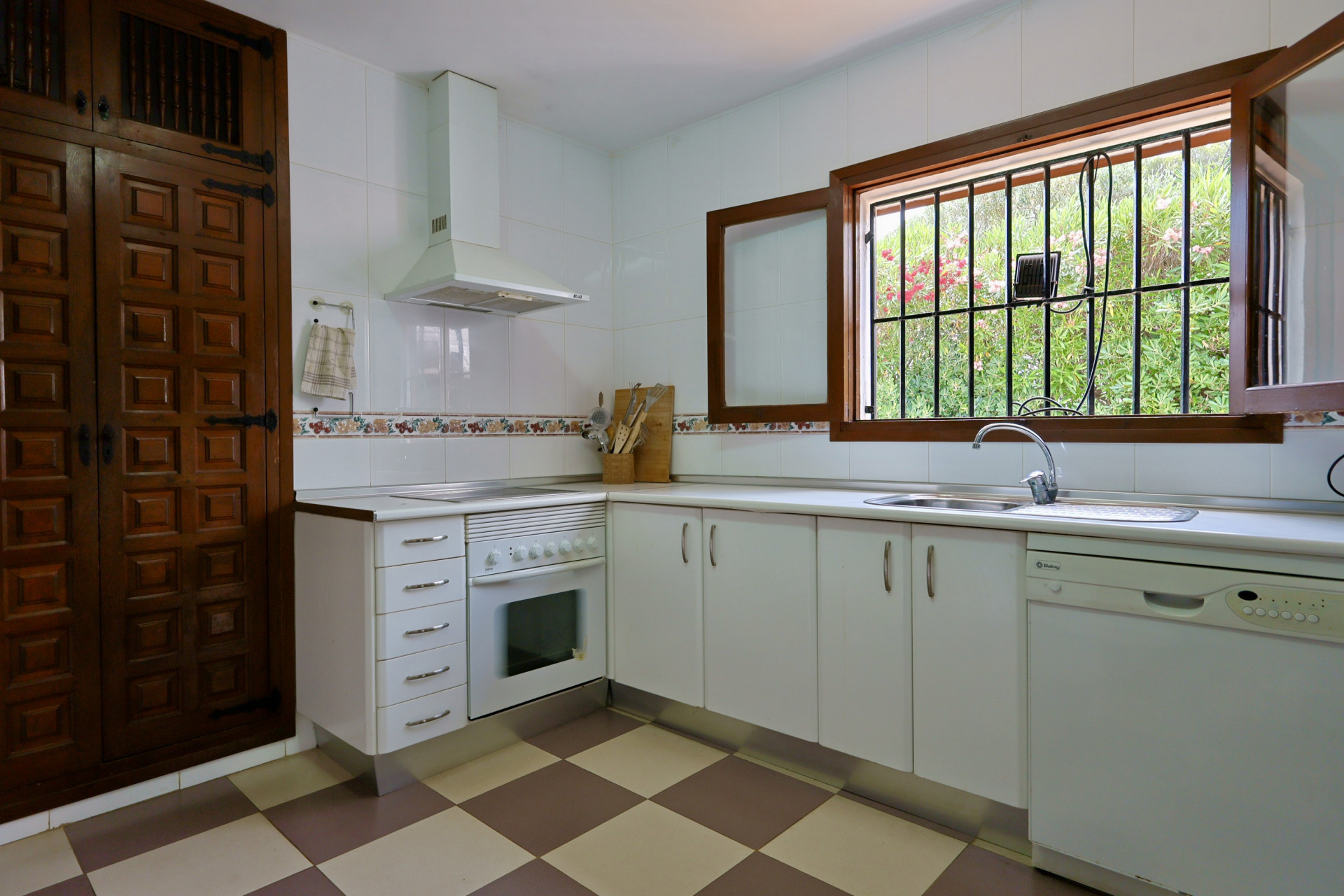 Functionele keuken met witte kasten, geblokte tegelvloer en een houten raam met tralies dat uitzicht biedt op de tuin. Voorzien van een RVS spoelbak en vaatwasser.