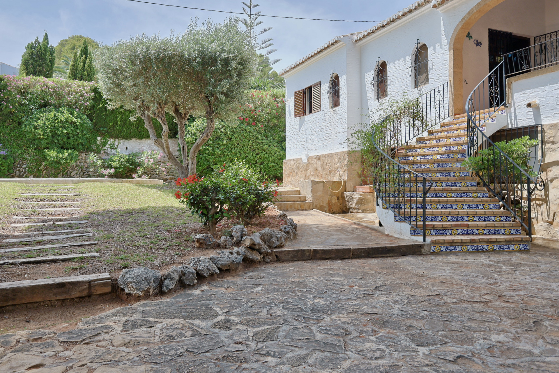 Hoofdingang van de villa met gebogen trap van decoratieve tegels en smeedijzeren balustrade, geflankeerd door een mediterrane tuin met olijfboom.