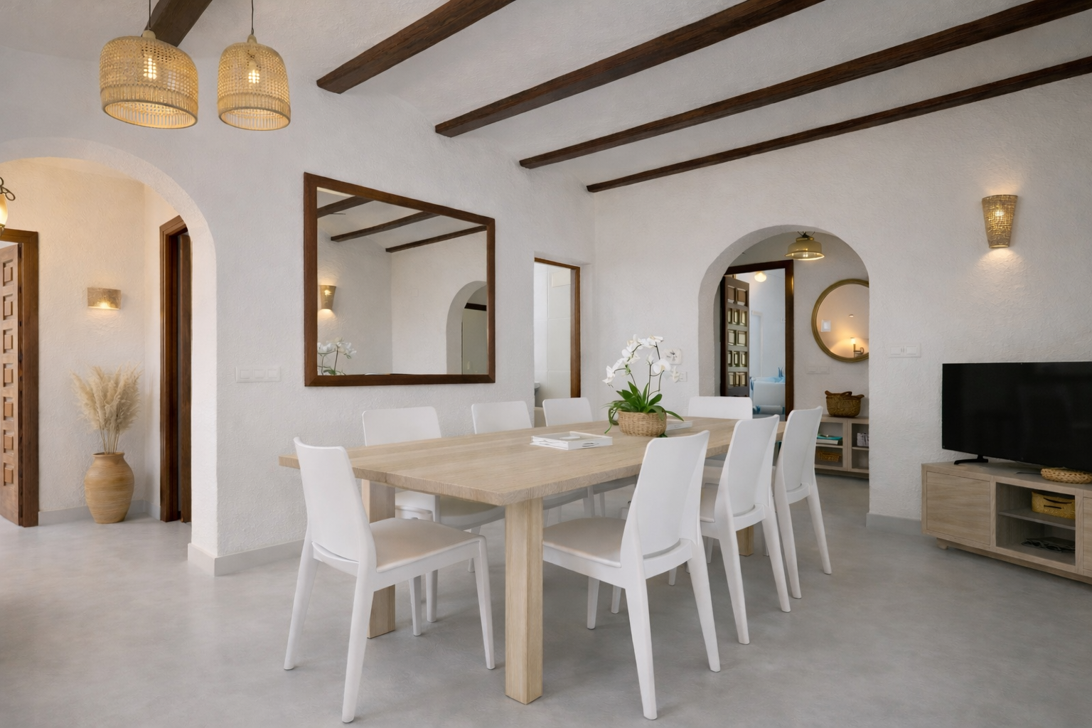 Lichte eetkamer met een lichte houten tafel voor acht en witte stoelen. Het plafond heeft donkere houten balken, rieten hanglampen en een grote spiegel met lijst.