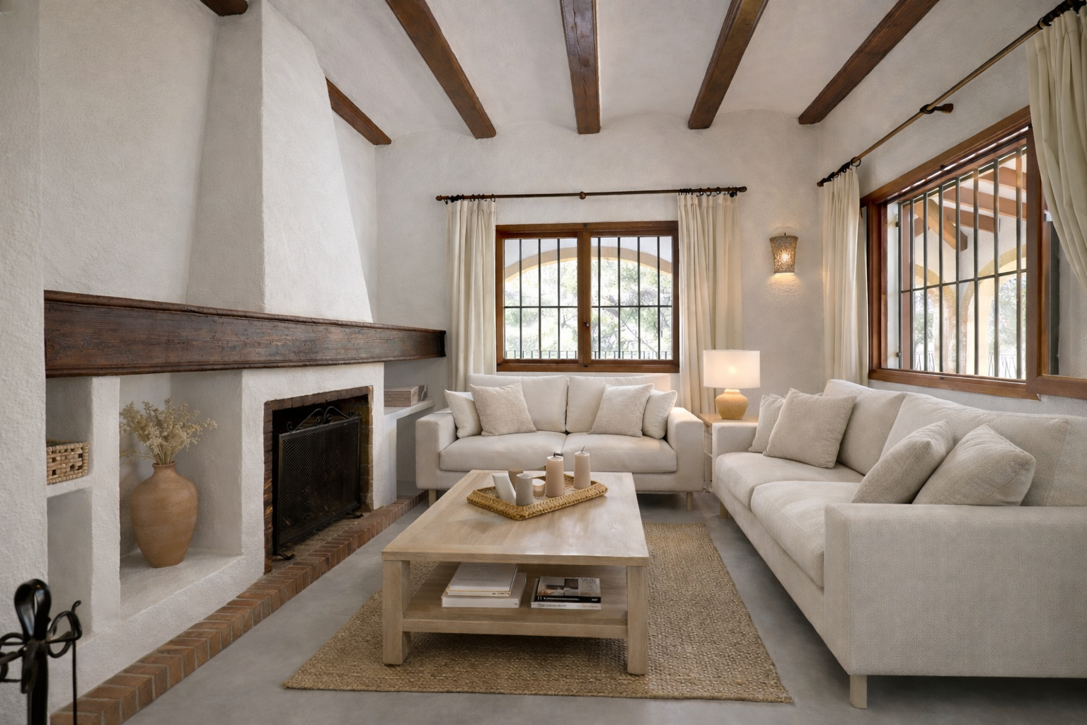 Woonkamer met traditionele witte stucwerk open haard en donkere houten schouw. Zichtbare plafondbalken en houten ramen met tralies kenmerken de ruimte.