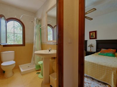 Badkamer met douche, toilet en wastafel op zuil, voorzien van een boogvormig houten raam met uitzicht op groen. Een glimp van een aangrenzende slaapkamer is zichtbaar.