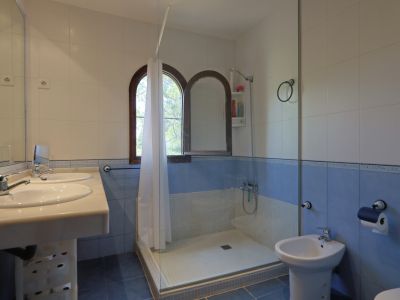 Badkamer met dubbele wastafel en licht aanrechtblad, wit-blauwe tegelwanden en inloopdouche met glazen scherm. Twee boogvormige houten ramen zorgen voor natuurlijk licht.