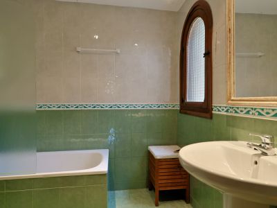 Badkamer met groen-beige tegels, ligbad met matglazen scherm en voetstukwastafel. Boograam van hout met gestructureerd glas biedt natuurlijk licht.