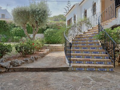 Buitentrap van baksteen met decoratieve blauw-gele tegels en smeedijzeren balustrade, geflankeerd door een mediterrane tuin met olijfboom.