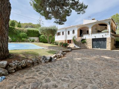 Buitenzijde van de villa met ruime geplaveide oprit, witte gevel met bogen en de hoofdtrap. Het afgedekte zwembad en mediterrane beplanting zijn zichtbaar.