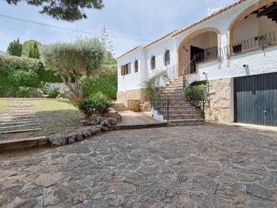 Exterieur van de villa met stenen oprit, een gebogen mozaïektrap en smeedijzeren balustrade. De mediterrane tuin omvat een olijfboom en een gazon naast de witte gevel.