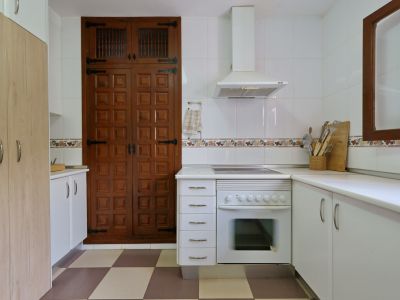 Functionele keuken met ingebouwde oven, afzuigkap en witte tegelwanden. Een opvallende grote houten kast met paneeldeuren en traditioneel beslag staat op een geblokte vloer.