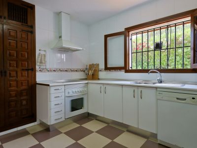 Functionele keuken met witte kasten, geblokte tegelvloer en een houten raam met tralies dat uitzicht biedt op de tuin. Voorzien van een RVS spoelbak en vaatwasser.
