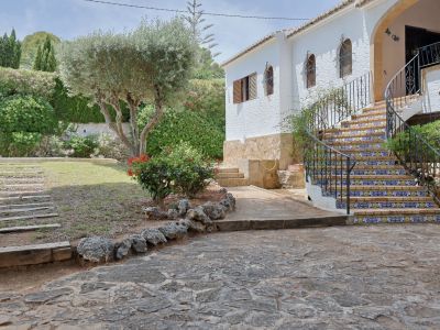 Hoofdingang van de villa met gebogen trap van decoratieve tegels en smeedijzeren balustrade, geflankeerd door een mediterrane tuin met olijfboom.