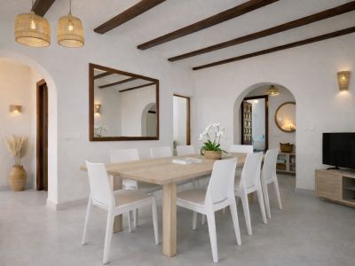 Lichte eetkamer met een lichte houten tafel voor acht en witte stoelen. Het plafond heeft donkere houten balken, rieten hanglampen en een grote spiegel met lijst.