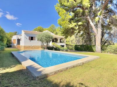 Rechthoekig zwembad met blauwe mozaïektegels, omgeven door een verzorgd groen gazon, met een witte villa met overdekt terras en mediterrane bomen op de achtergrond.