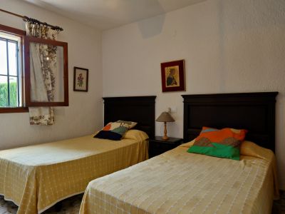 Tweepersoons slaapkamer met donkere houten hoofdeinden, gele sprei met patroon en kleurrijke kussens. Raam met tralies en bloemengordijnen.
