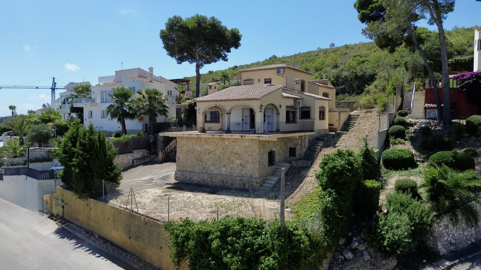 Buitenaanzicht van een mediterrane villa met twee verdiepingen, stenen basis en gewelfd balkon. Gelegen op hellend terrein met vegetatie onder een heldere blauwe lucht.