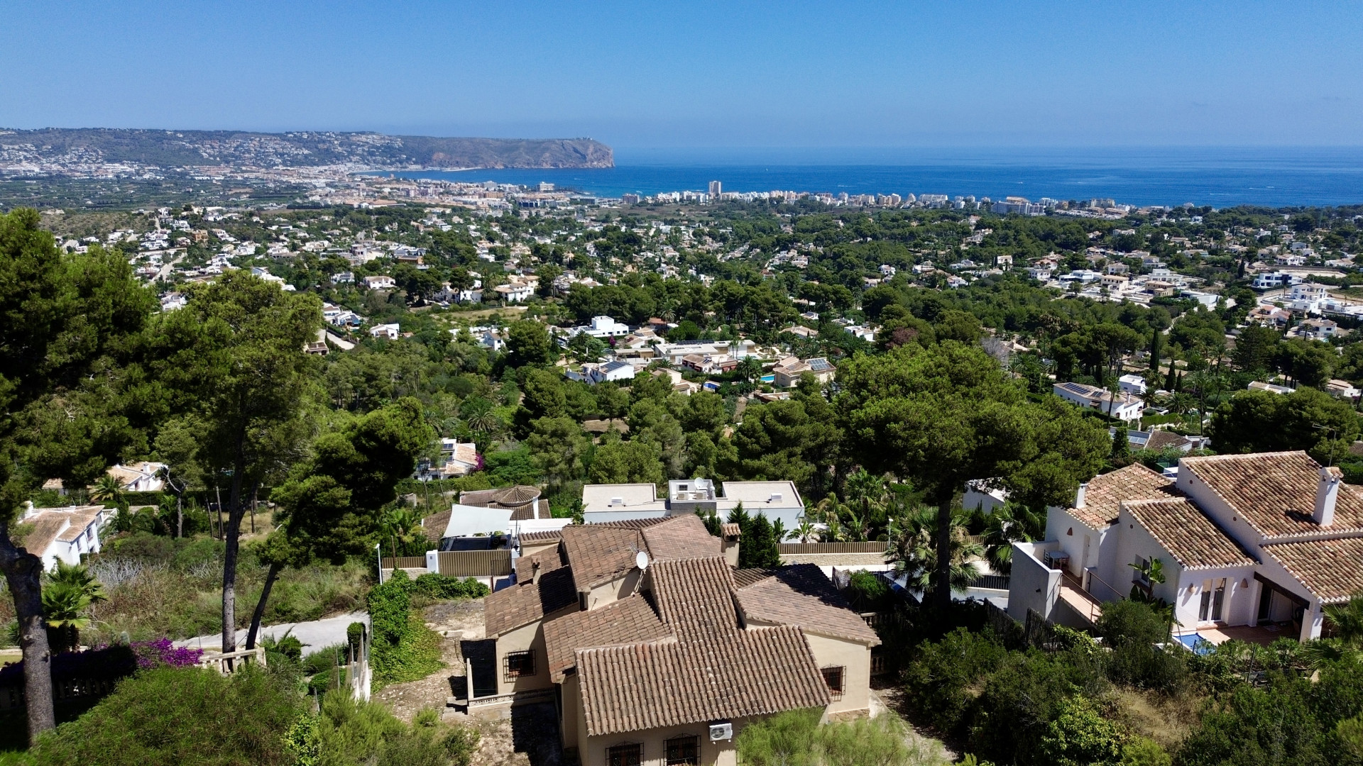Luchtfoto van Jávea, met traditionele villa's met terracotta daken tussen pijnbomen, en de Middellandse Zee en Cabo de San Antonio op de achtergrond.