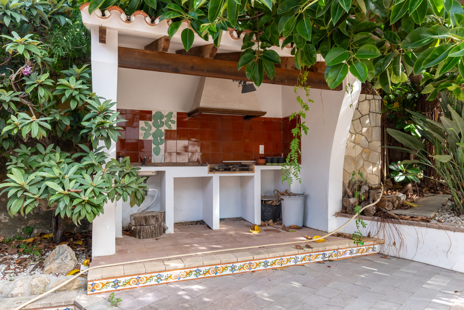 Buitenkeuken met ingebouwde BBQ, terracotta tegelachterwand met cactusmotief en rustieke houten balkenplafond. Het gebied is betegeld en omgeven door weelderig groen.