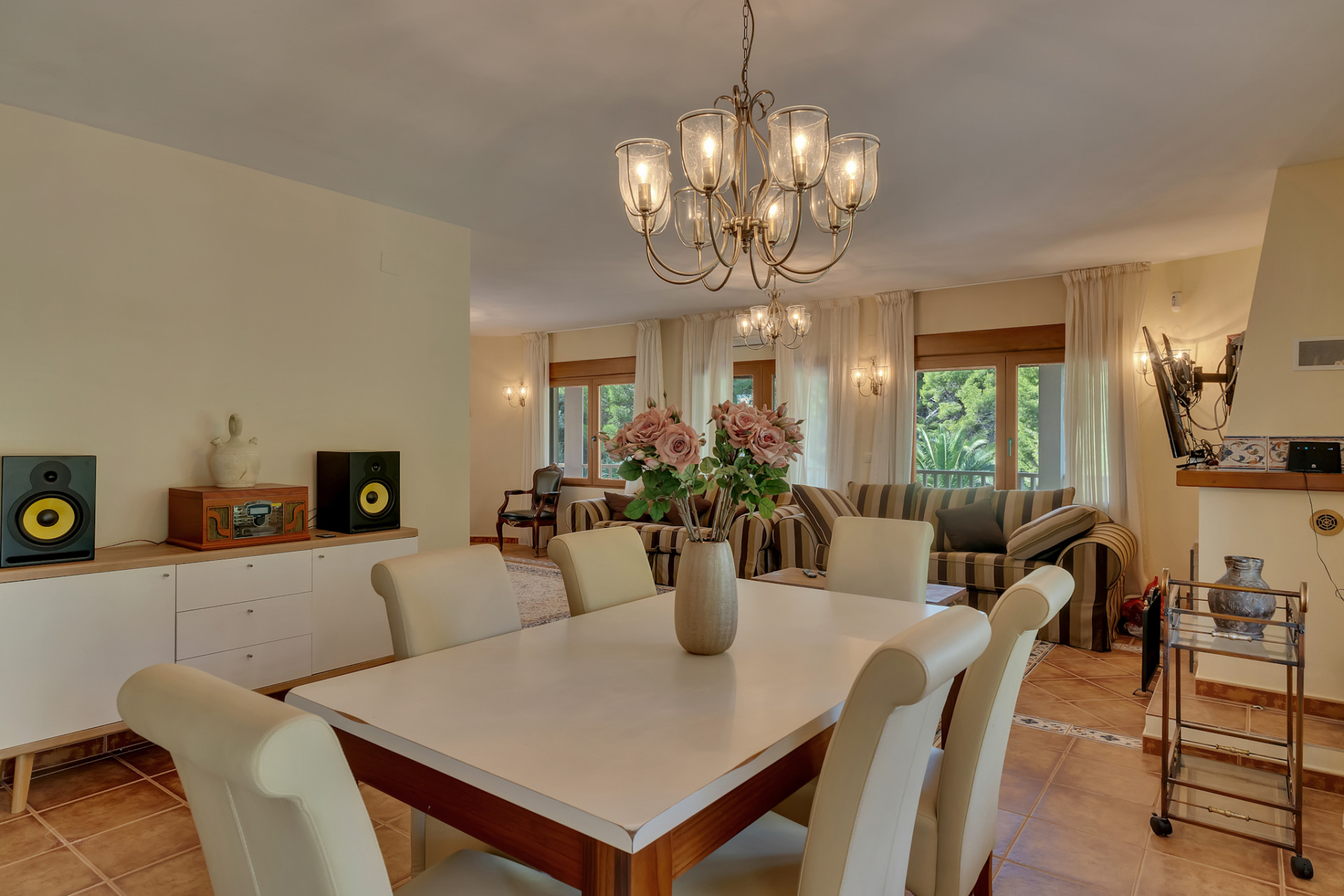 Elegante eetruimte met klassieke kroonluchter en crèmekleurige stoelen, naadloos verbonden met een lichte woonkamer met gestreepte banken en grote ramen.