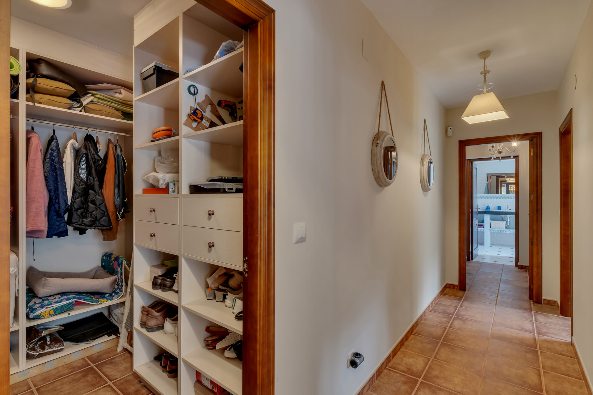 Gang met geïntegreerde open kledingkast, witte planken en lades, terracotta vloertegels en houten deurkozijnen.