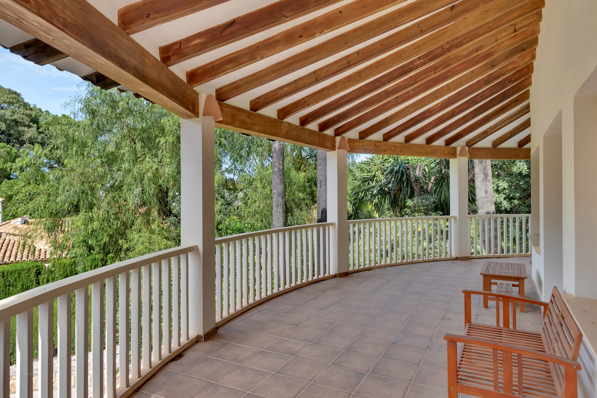 Gebogen overdekt terras met tegelvloer en een plafond van zichtbare houten balken. De witte balustrade biedt uitzicht op weelderig groen, aangevuld met eenvoudige houten zitbanken.