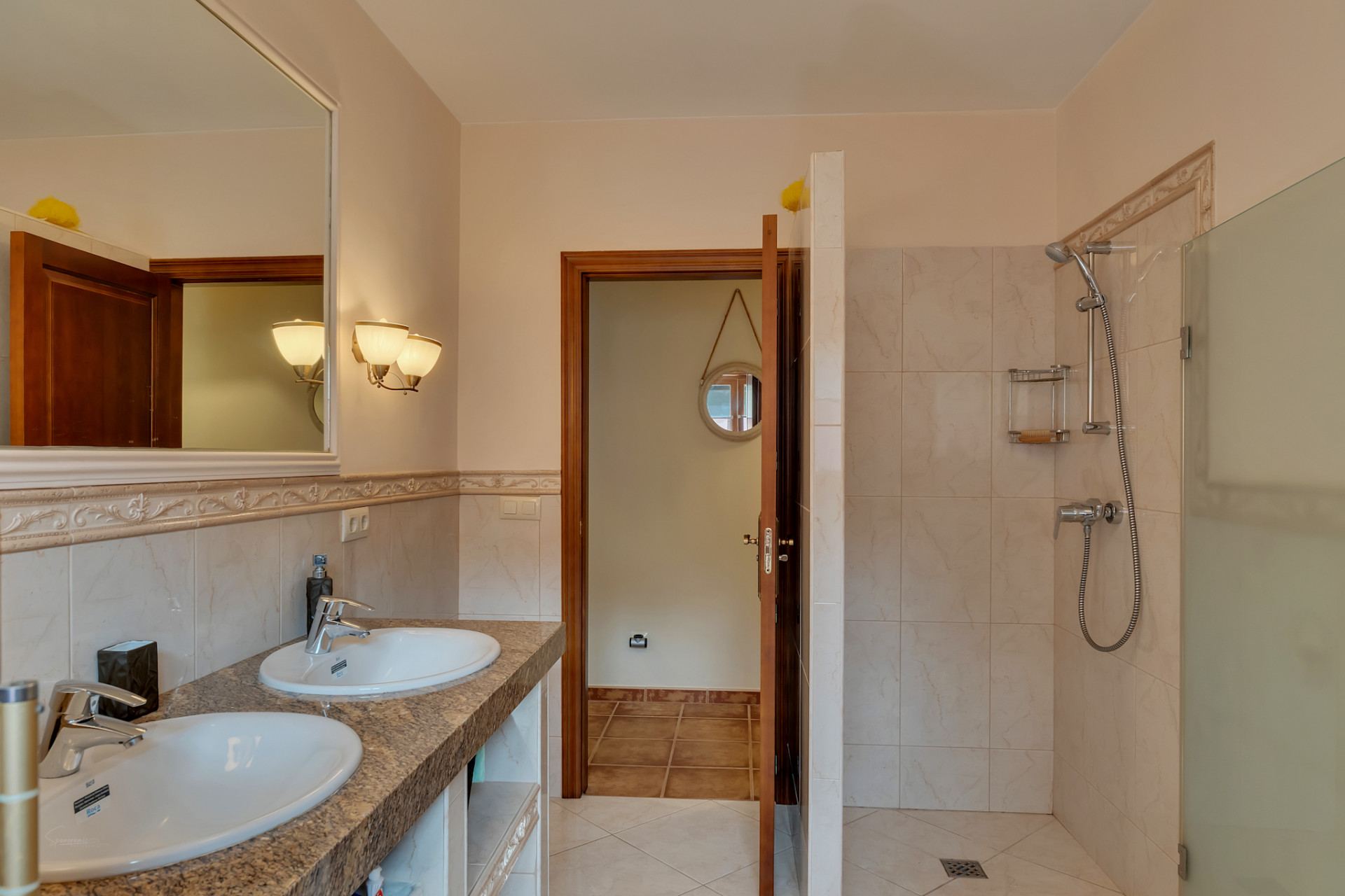 Hoofdbadkamer met dubbele wastafel op granieten aanrecht, grote ingelijste spiegel en inloopdouche met glazen wand, geaccentueerd door warm getinte tegels.