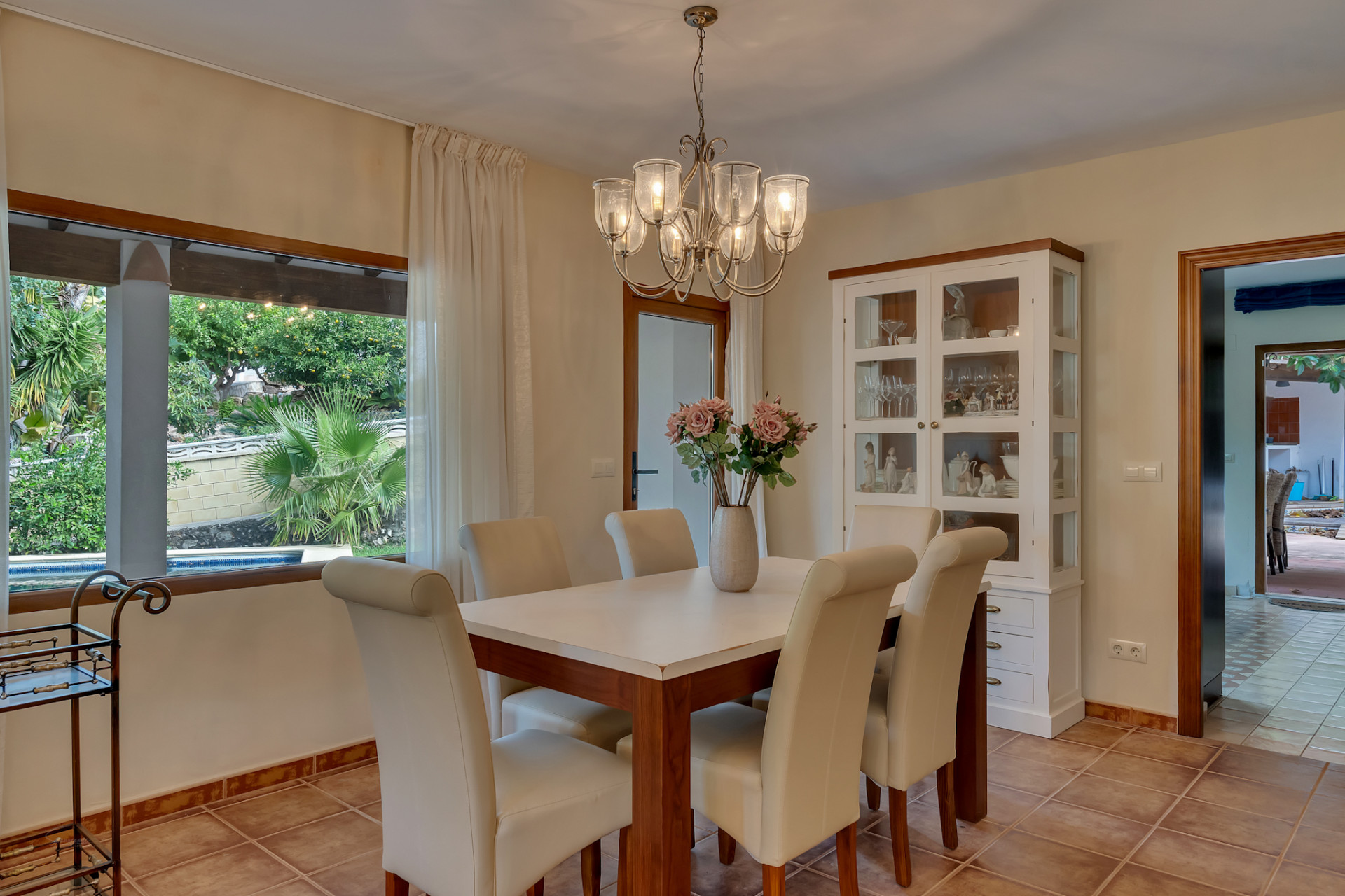 Lichte eetkamer met houten tafel, crèmekleurige gestoffeerde stoelen en kroonluchter. Groot raam biedt uitzicht op de weelderige tuin en het privézwembad.