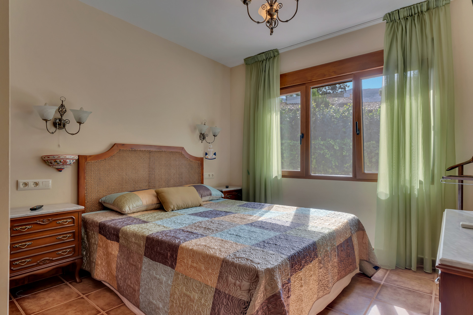Lichte slaapkamer met rotan hoofdbord, een patchwork sprei en een groot houten raam met lichtgroene gordijnen. Terracottategels en klassieke houten nachtkastjes.