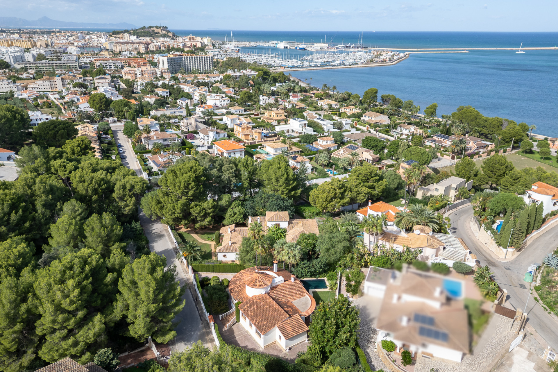 Luchtfoto van de villa met rood pannendak en privézwembad, gelegen in een groene woonwijk van Dénia, met uitzicht op de jachthaven en de zee.