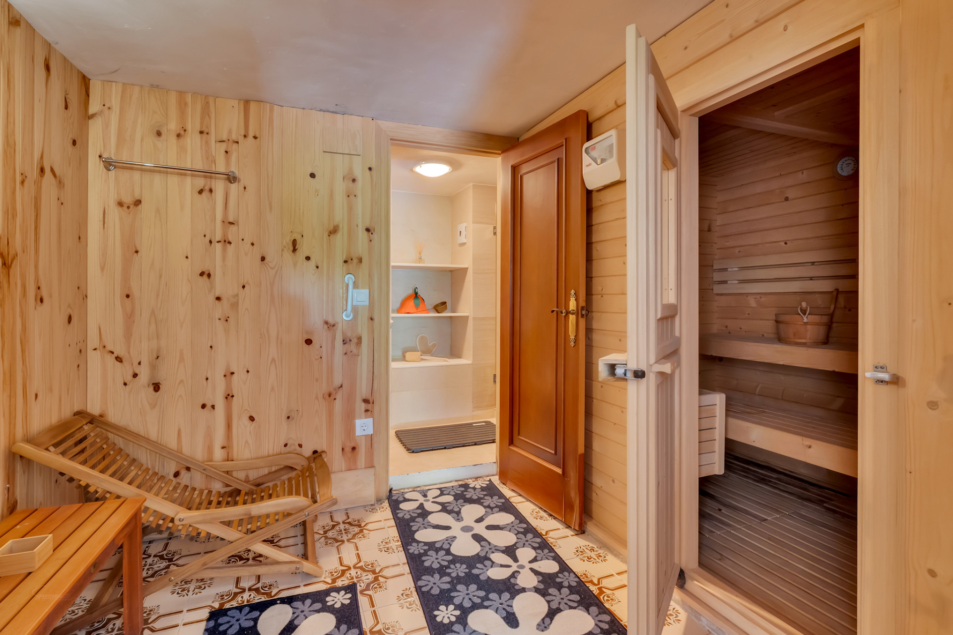 Ontspanningsruimte met lichte houten wanden en tegelvloer, direct toegang tot een grenenhouten sauna. Een houten ligstoel en een aangrenzende opbergruimte met planken.