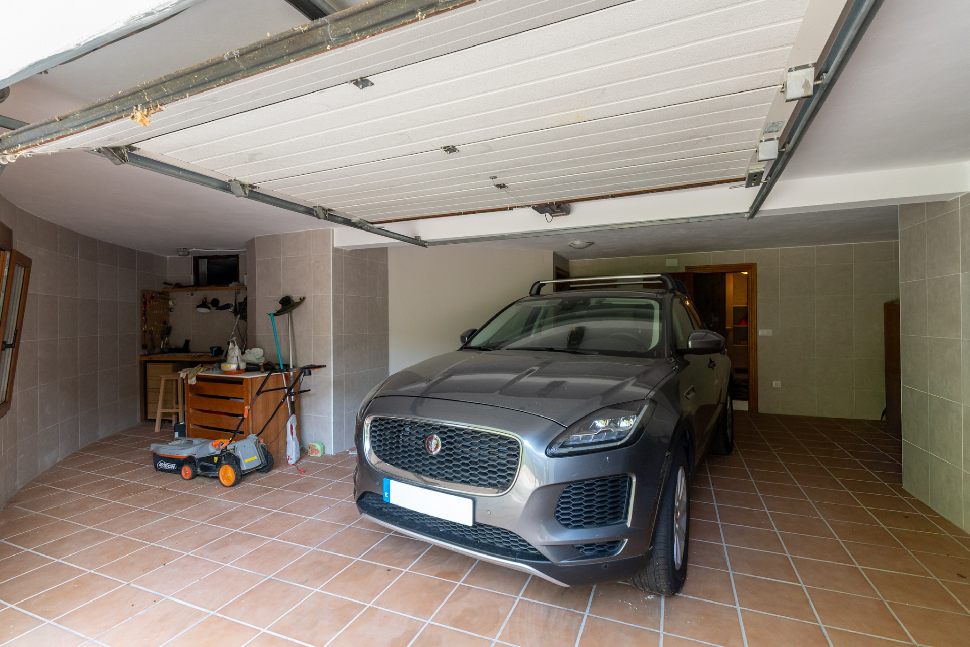 Privégarage met terracotta tegelvloer en grijze wandtegels. De ruimte omvat een werkbank met gereedschap, een grasmaaier en een open sectionale garagedeur.
