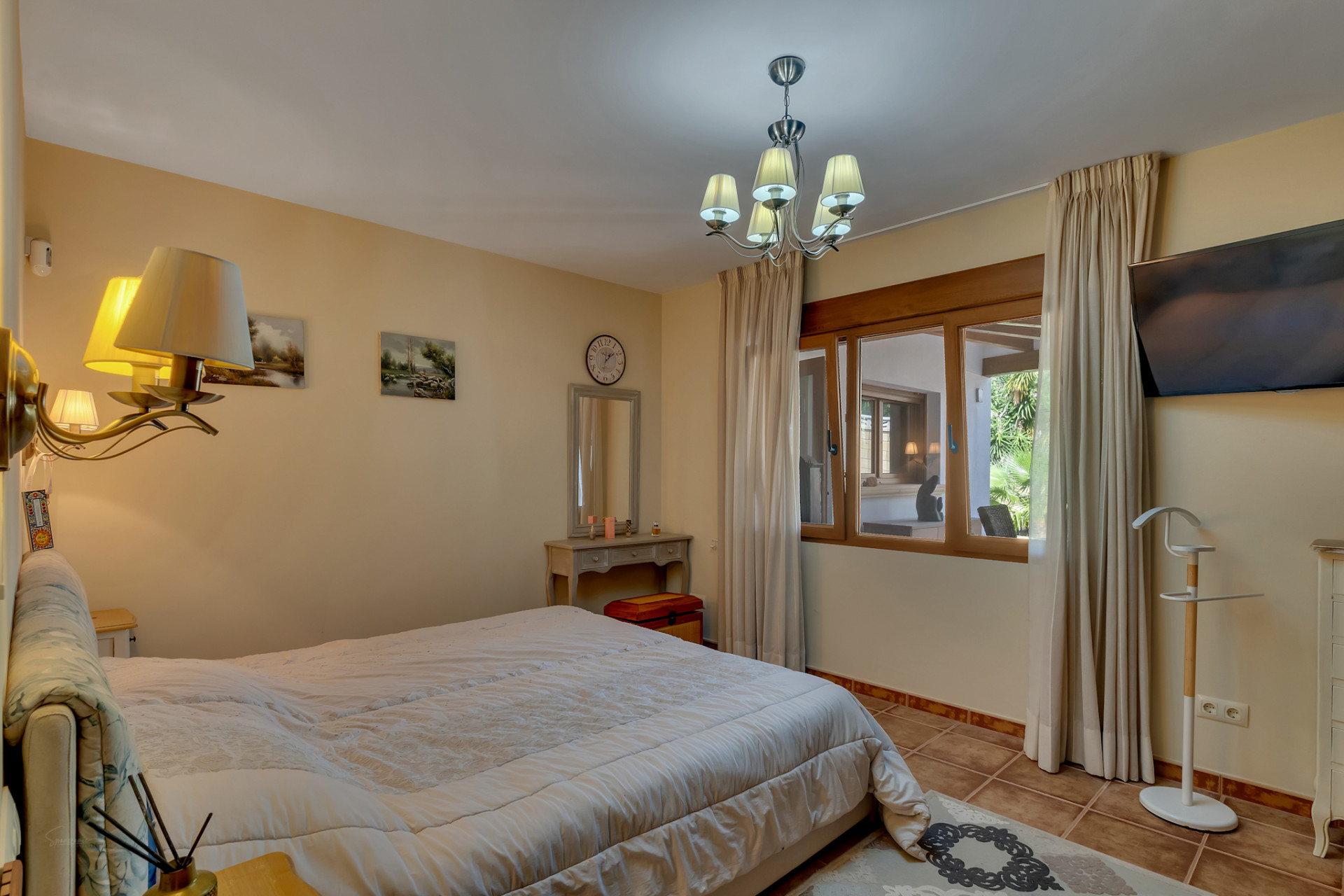 Ruime slaapkamer met groot bed, tegelvloer en houten raamkozijn met uitzicht op een aangrenzende overdekte ruimte en tuin. Voorzien van een kaptafel en wandtelevisie.