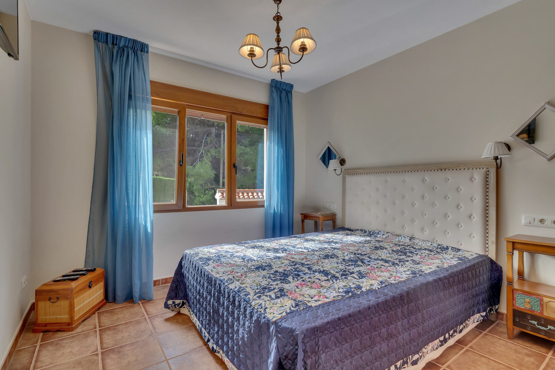 Slaapkamer met groot bed, wit gestoffeerd hoofdbord en blauw gedessineerde sprei. Terracotta tegelvloer en houten raam met blauwe gordijnen bieden uitzicht op groen.