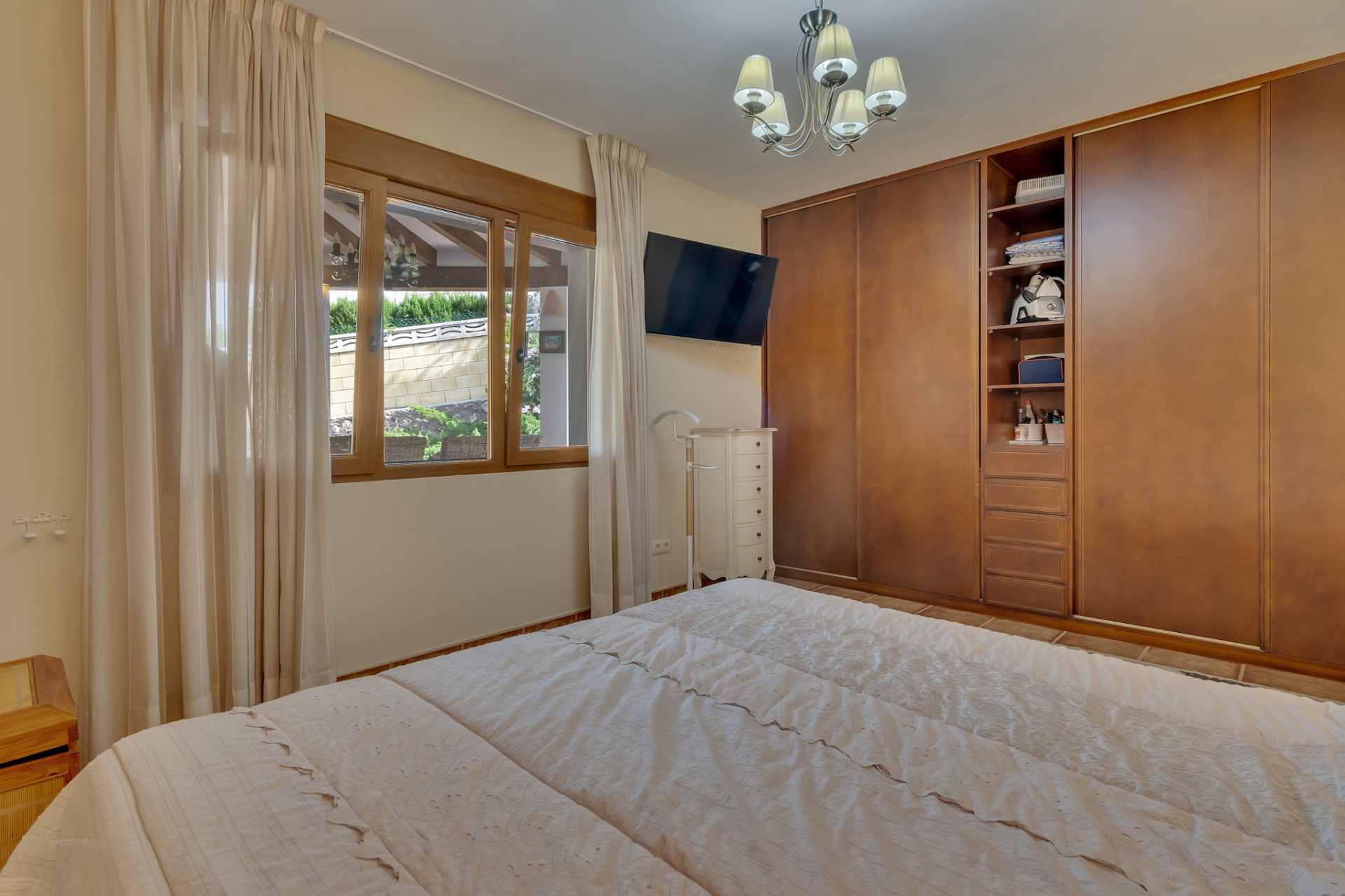 Slaapkamer met tweepersoonsbed, ingebouwde houten kasten en raam met uitzicht op een aangelegde muur. Voorzien van een wand-tv en witte ladekast.