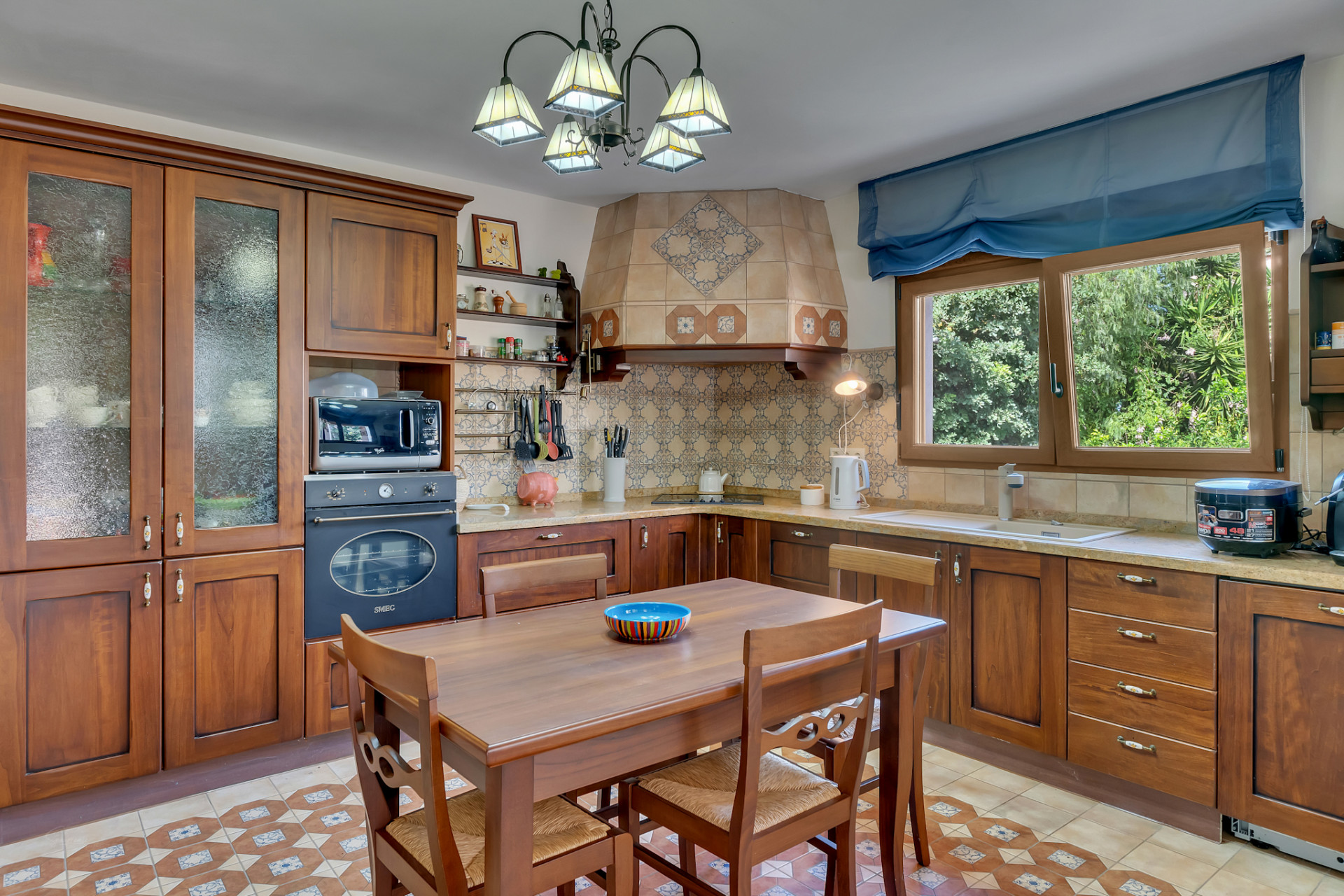 Traditionele keuken met donkerhouten kasten, een betegelde achterwand met patronen en een centrale eettafel. Natuurlijk licht stroomt binnen via het raam met uitzicht op groen.