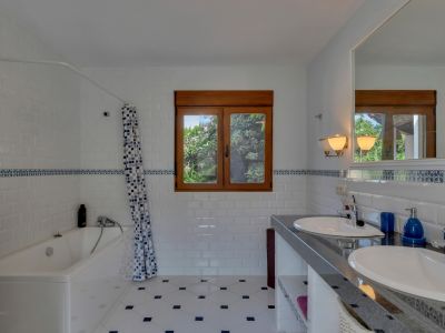 Badkamer met dubbele wastafel op donker aanrechtblad en grote spiegel. Witte metrotegels met blauwe sierrand, patroonvloer en houten raam met uitzicht op groen.