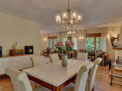 Elegante eetruimte met klassieke kroonluchter en crèmekleurige stoelen, naadloos verbonden met een lichte woonkamer met gestreepte banken en grote ramen.