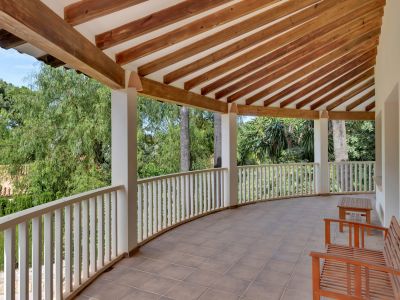 Gebogen overdekt terras met tegelvloer en een plafond van zichtbare houten balken. De witte balustrade biedt uitzicht op weelderig groen, aangevuld met eenvoudige houten zitbanken.
