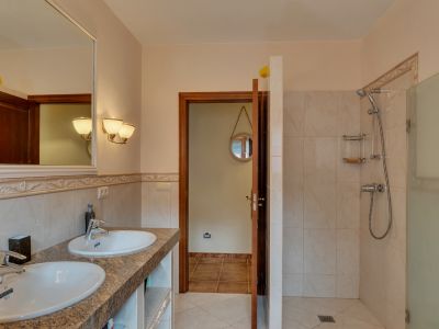 Hoofdbadkamer met dubbele wastafel op granieten aanrecht, grote ingelijste spiegel en inloopdouche met glazen wand, geaccentueerd door warm getinte tegels.