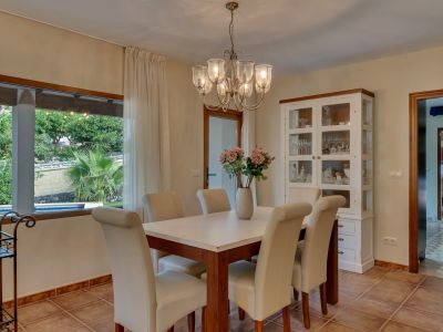 Lichte eetkamer met houten tafel, crèmekleurige gestoffeerde stoelen en kroonluchter. Groot raam biedt uitzicht op de weelderige tuin en het privézwembad.