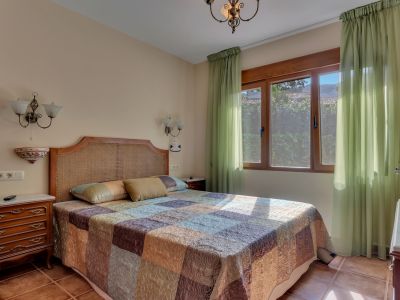 Lichte slaapkamer met rotan hoofdbord, een patchwork sprei en een groot houten raam met lichtgroene gordijnen. Terracottategels en klassieke houten nachtkastjes.