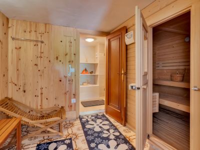 Ontspanningsruimte met lichte houten wanden en tegelvloer, direct toegang tot een grenenhouten sauna. Een houten ligstoel en een aangrenzende opbergruimte met planken.