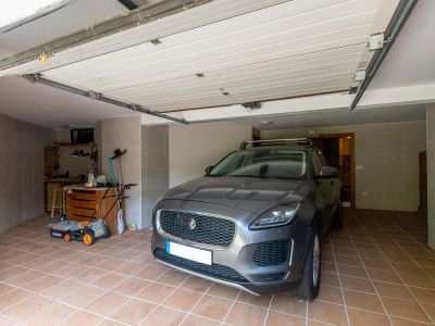 Privégarage met terracotta tegelvloer en grijze wandtegels. De ruimte omvat een werkbank met gereedschap, een grasmaaier en een open sectionale garagedeur.