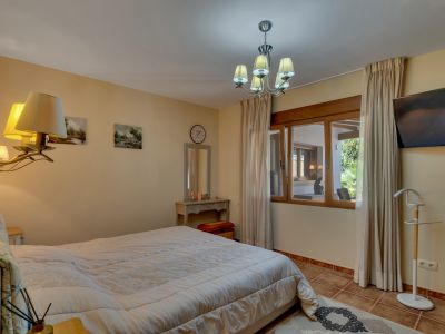 Ruime slaapkamer met groot bed, tegelvloer en houten raamkozijn met uitzicht op een aangrenzende overdekte ruimte en tuin. Voorzien van een kaptafel en wandtelevisie.